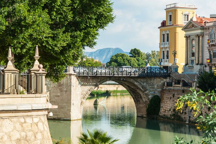 Eine alte Steinbrücke mit Bögen führt über einen ruhigen Fluss, gesäumt von grünen Bäumen und ockerfarbenen historischen Gebäuden unter blauem Himmel.