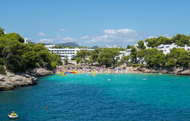 Viele Menschen baden und entspannen an einer kleinen Strandbucht mit türkisfarbenem Wasser und weißen Gebäuden im Hintergrund.