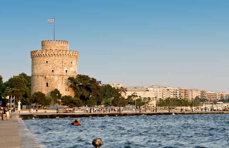 Ein runder Steinturm mit griechischer Flagge steht direkt an der Promenade am Wasser.