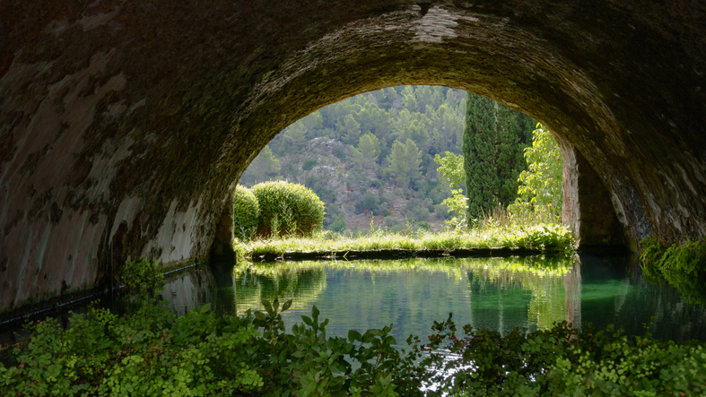 Ein stilles Wasserbecken liegt unter einem dunklen Torbogen mit Blick ins Grüne.