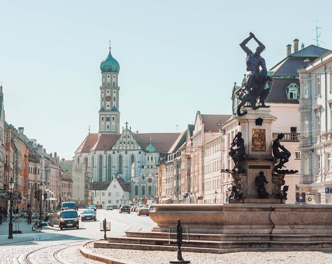 Der prunkvolle Herkulesbrunnen in einer Straßenszene in Augsburg mit der Basilika St. Ulrich und Afra im Hintergrund.