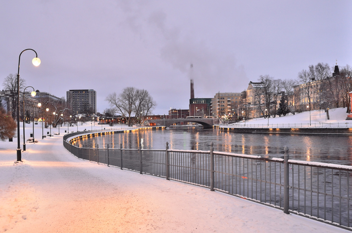 Ein verschneiter Uferweg mit Laternen verläuft an einem ruhigen Wasserlauf durch die Stadt.
