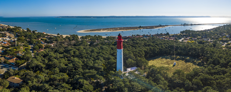 Ein rotweißer Leuchtturm ragt über einen bewaldeten Küstenstreifen mit Wasser und Sandflächen hinaus.