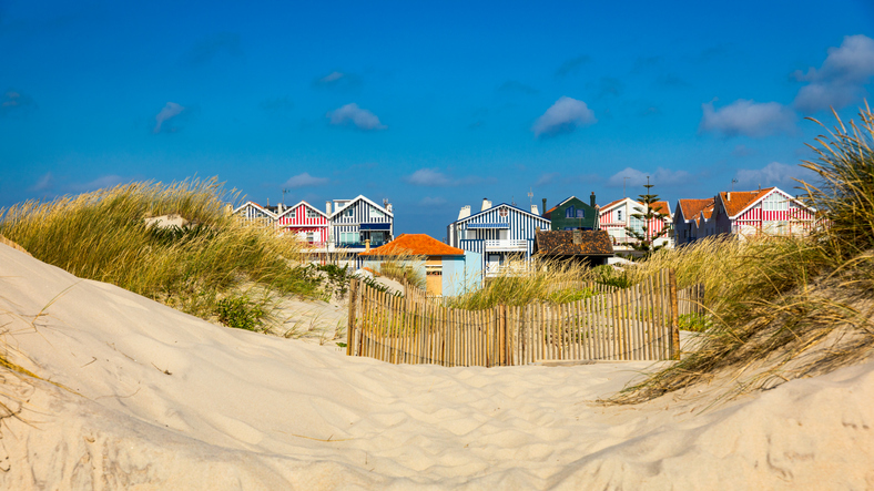 Farbenfrohe Strandhäuser hinter Dünen und einem Holzzaun unter blauem Himmel.