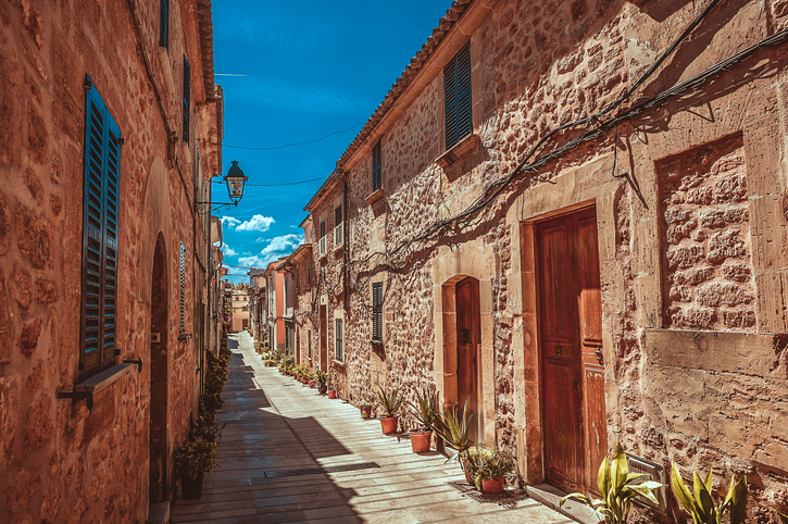 Eine schmale, sonnige Gasse in Alcudia mit rustikalen Steinfassaden, grünen Fensterläden und Palmen unter blauem Himmel.