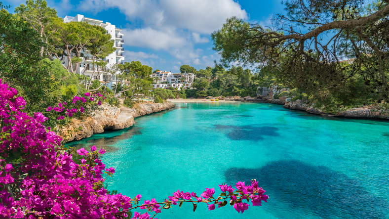 Eine ruhige Bucht mit leuchtend türkisem Wasser, Felsen, Bäumen und pinken Bougainvillea liegt unter blauem Himmel.