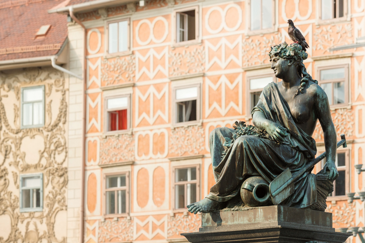 Detailaufnahme einer bronzenen Frauenstatue vor einer kunstvoll verzierten, hellen Hausfassade in Graz.