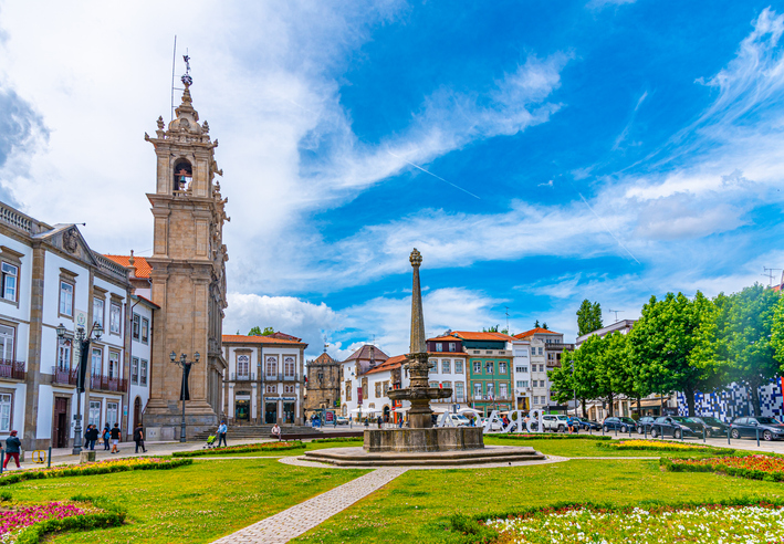 Ein sonniger Platz mit einem Brunnen und Blumenbeeten vor historischen Gebäuden und Kirchen in Braga.