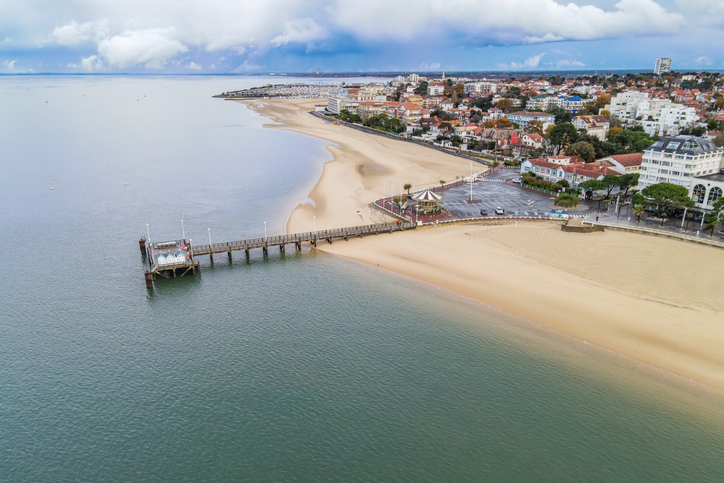 Ein langer Sandstrand, ein Steg und die Küstenbebauung ziehen sich unter wolkigem Himmel am Wasser entlang.