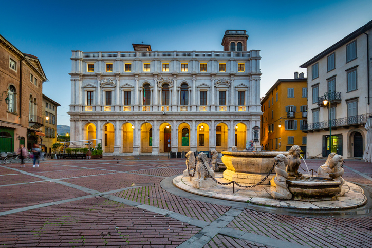 Ein heller Platz in Bergamo wird von einem Brunnen und einem prächtigen historischen Gebäude mit Arkaden geprägt.