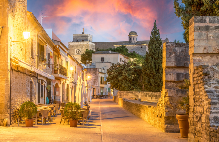 Eine beleuchtete Gasse in Alcudia während der Dämmerung mit Außengastronomie und der Silhouette der Kirche im Hintergrund.