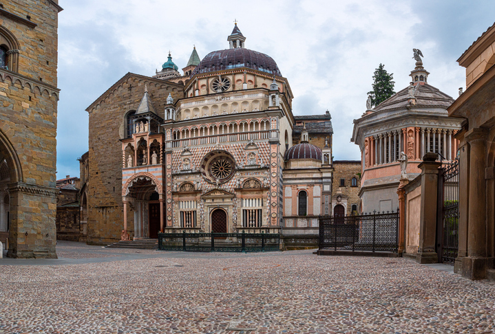 Eine reich verzierte Kirchenfassade mit Kuppeln und steinernem Vorplatz steht zwischen alten Gebäuden in Bergamo.