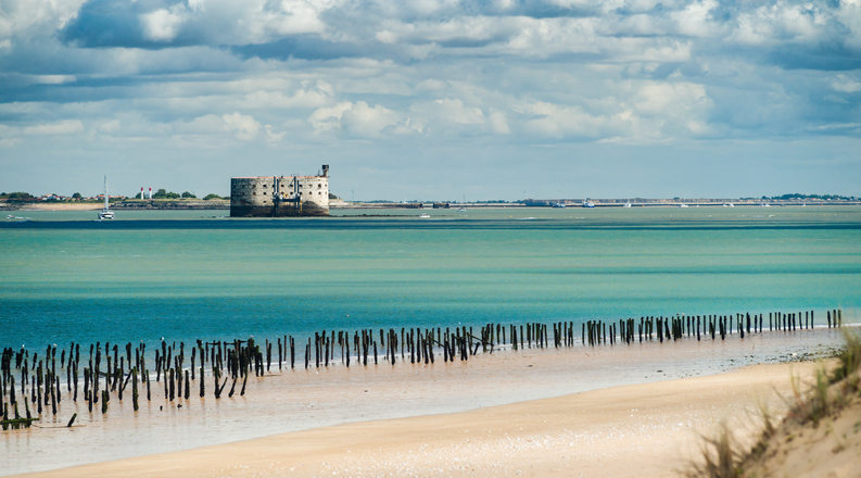 Sandstrand mit Holzpfählen im Vordergrund und dem fernen Fort Boyard am Horizont unter einem dramatischen Wolkenhimmel.