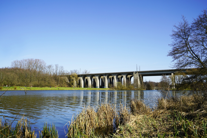 Eine lange Brücke mit Bögen spannt sich über einen ruhigen Fluss, an dessen Ufer Schilf und Bäume stehen.
