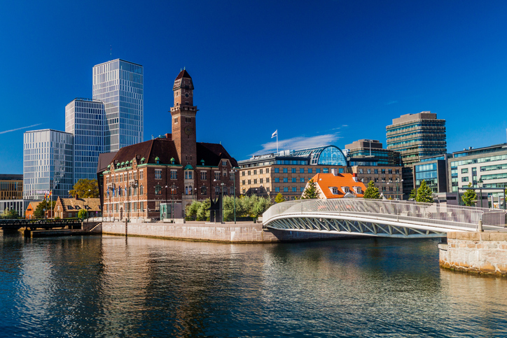 Blick über das Wasser auf das historische Backsteingebäude der Universität Malmö, kontrastiert durch moderne Hochhäuser im Hintergrund.