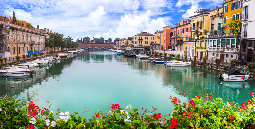 Ein blauer Kanal mit zahlreichen Booten, gesäumt von bunten Häuserfassaden und Blumen in Peschiera del Garda am Gardasee.