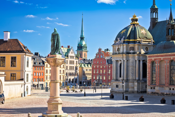 Eine Statue auf einer hohen Säule blickt über den sonnigen Platz auf die prachtvollen Kuppeln und Giebel der historischen Gebäude.