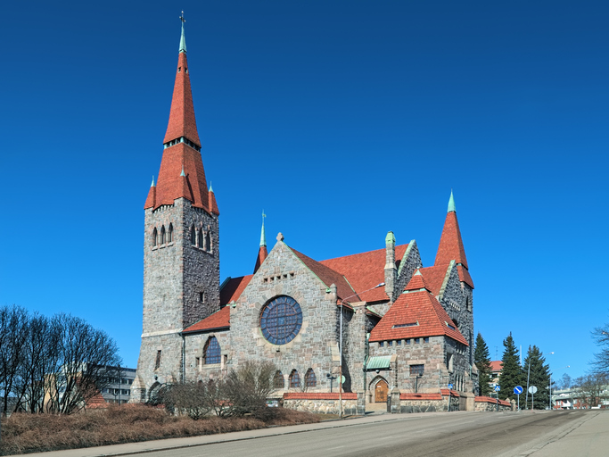 Eine große Kirche aus grauem Stein mit roten Dächern und hohen Türmen steht an einer breiten Straße.