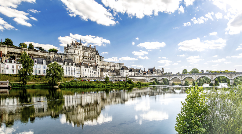 Ein großes Schloss, eine steinerne Brücke und historische Häuser spiegeln sich in einem ruhigen Fluss.
