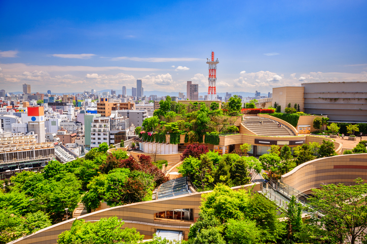 Eine moderne Dachlandschaft mit viel Grün, Terrassen und roten Sendemasten liegt über der Skyline von Osaka.