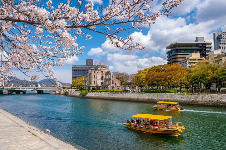 Kirschblüten rahmen einen Fluss in Hiroshima, auf dem gelbe Boote fahren, während am Ufer ein markantes historisches Gebäude steht.