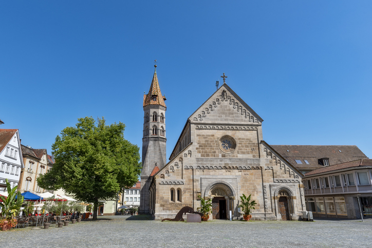 Eine helle Kirche mit hohem Turm steht auf einem ruhigen Platz unter blauem Himmel.