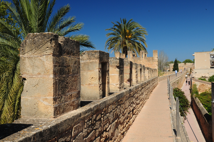 Ein gepflasterter Wehrgang oben auf der historischen Stadtmauer von Alcudia mit Blick auf Palmen und die Stadt.