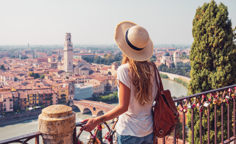 Eine Touristin mit Strohhut blickt von einer Aussichtsplattform mit Liebesschlössern über das Panorama der Altstadt von Verona.