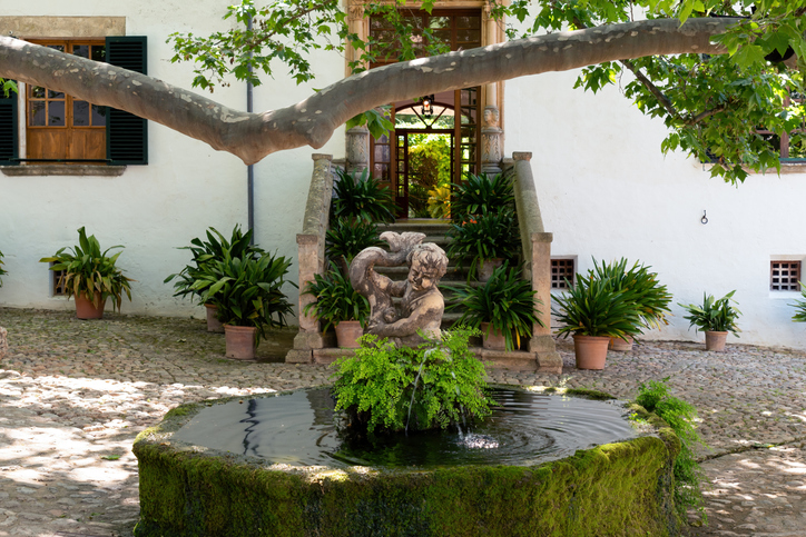 Ein moosiger Brunnen mit Figur steht vor einem hellen Gebäude in einem Innenhof.