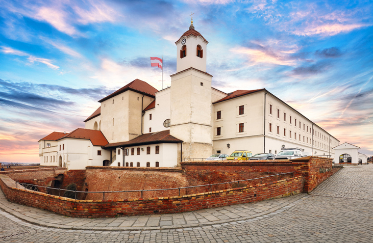 Eine helle Festungsanlage steht auf einer Mauer unter farbigem Himmel.
