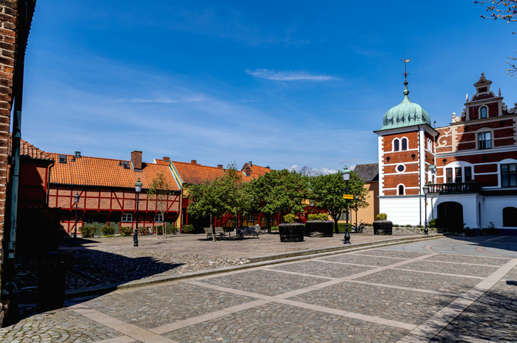 Ein weiter, gepflasterter Platz mit Bäumen, umgeben von historischen Backsteinbauten und einem markanten Turmgebäude.
