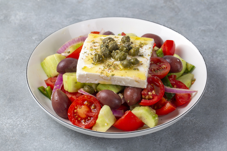 Eine weiße Schüssel mit frischem griechischem Salat, getoppt mit einem großen Block Feta-Käse, Oliven und Kapern.