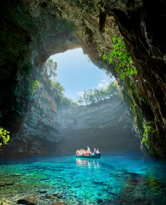 Ein Boot mit Passagieren gleitet über spiegelglattes, azurblaues Wasser in einer unterirdischen Höhle, in die Sonnenlicht von oben einfällt.