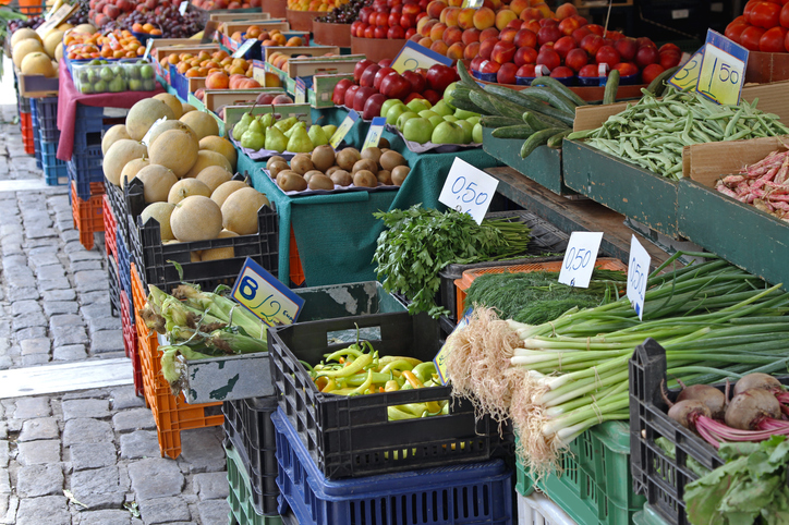 Kisten mit Gemüse, Obst und Kräutern stehen dicht nebeneinander an einem Marktstand.