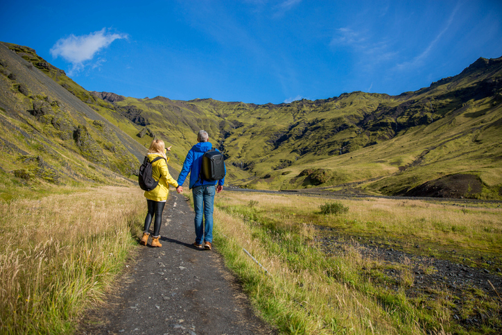 Ein Paar wandert Hand in Hand auf einem Pfad durch eine grüne Hügellandschaft mit Bergen im Hintergrund.