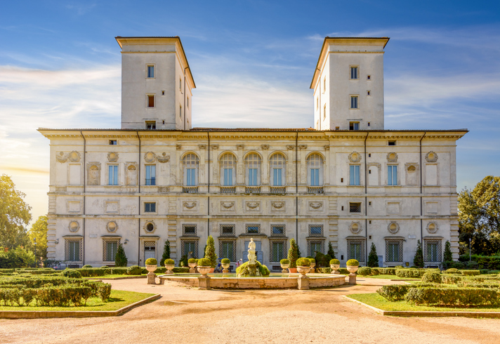 Die prachtvolle Fassade der Galleria Borghese mit zwei Türmen, davor ein gepflegter Garten mit Brunnen und Skulptur.