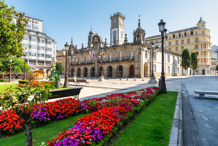 Das barocke Rathaus von Lugo mit seinem markanten Uhrturm, davor farbenfrohe Blumenbeete auf dem belebten Hauptplatz.