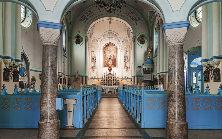 Der Innenraum einer Kirche zeigt helle Säulen, blaue Bänke und einen reich verzierten Altar.