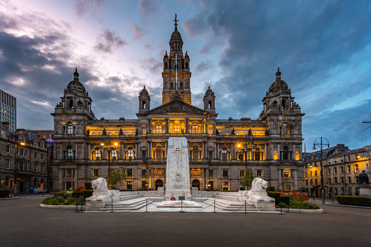 Die beleuchtete Prachtfassade des Rathauses von Glasgow am George Square unter einem dramatischen Abendhimmel.
