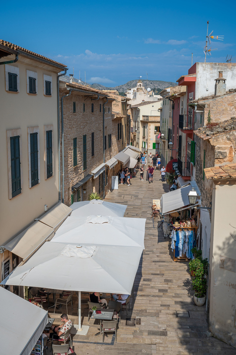 Eine belebte Fußgängerzone in der Altstadt von Alcudia mit Cafés unter weißen Sonnenschirmen und Blick auf die Berge.