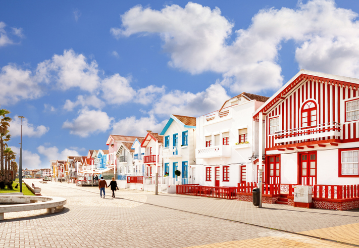 Spaziergänger auf der gepflasterten Straße vor weißen und rot gestreiften Häusern unter blauem Himmel.