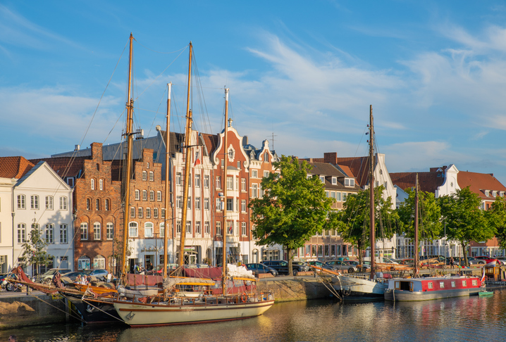 Alte Segelschiffe mit hohen Masten liegen an der Kaimauer vor einer Reihe prächtiger Giebelhäuser bei strahlendem Sonnenschein.