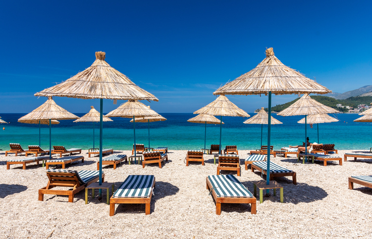 Mehrere strohgedeckte Sonnenschirme und Holzliegen stehen auf einem hellen Strand vor türkisfarbenem Wasser.