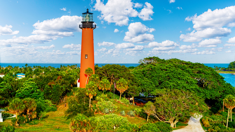 Ein hoher orangefarbener Leuchtturm ragt über dichte grüne Bäume unter wolkigem Himmel.