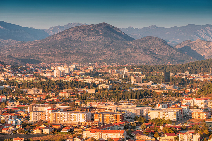Eine weite Stadtansicht liegt vor einer großen Berglandschaft im warmen Licht.