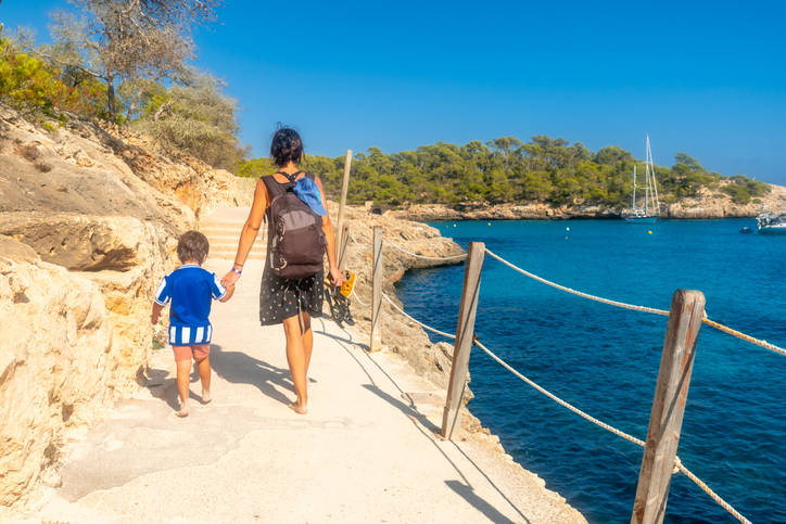 Eine Frau und ein kleines Kind gehen Hand in Hand einen hellen Küstenpfad entlang, der an einer türkisfarbenen Bucht mit Segelbooten vorbeiführt.
