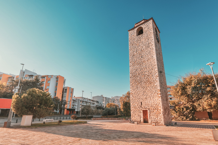 Ein schmaler Steinturm steht auf einem offenen Platz unter blauem Himmel.
