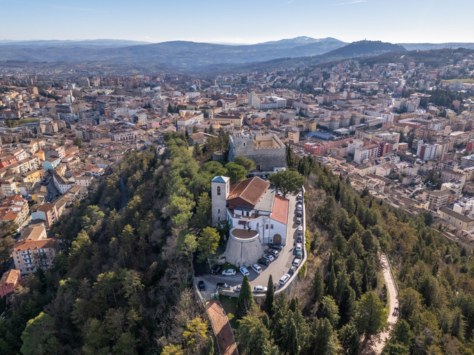 Luftbild einer weißen Kirche mit rotem Dach auf einem bewaldeten Hügel über der Stadt mit Bergpanorama.