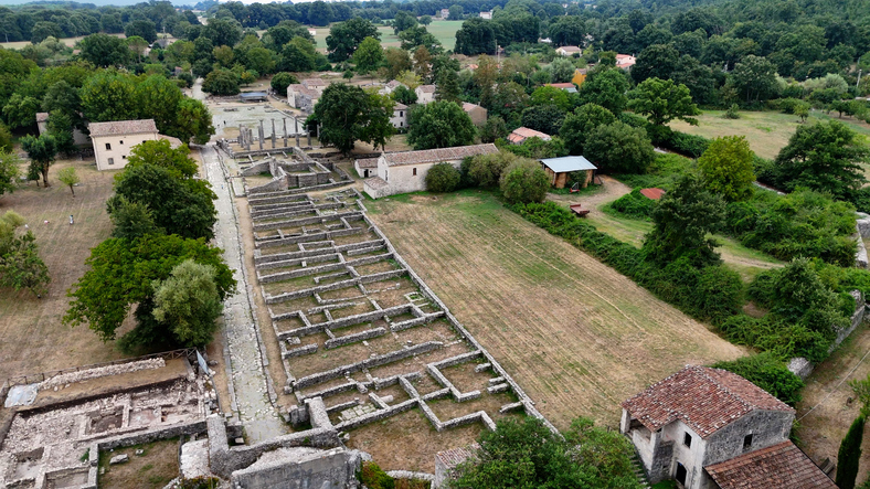 Luftaufnahme großer antiker Ruinen mit Grundmauern inmitten grüner Landschaft und alter Häuser.