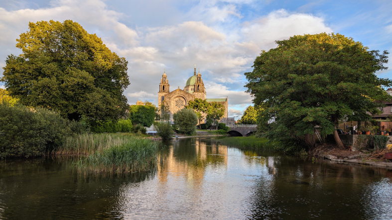 Die massive Steinkirche mit ihrer grünen Kuppel spiegelt sich in einem ruhigen Kanalarm des Flusses Corrib.